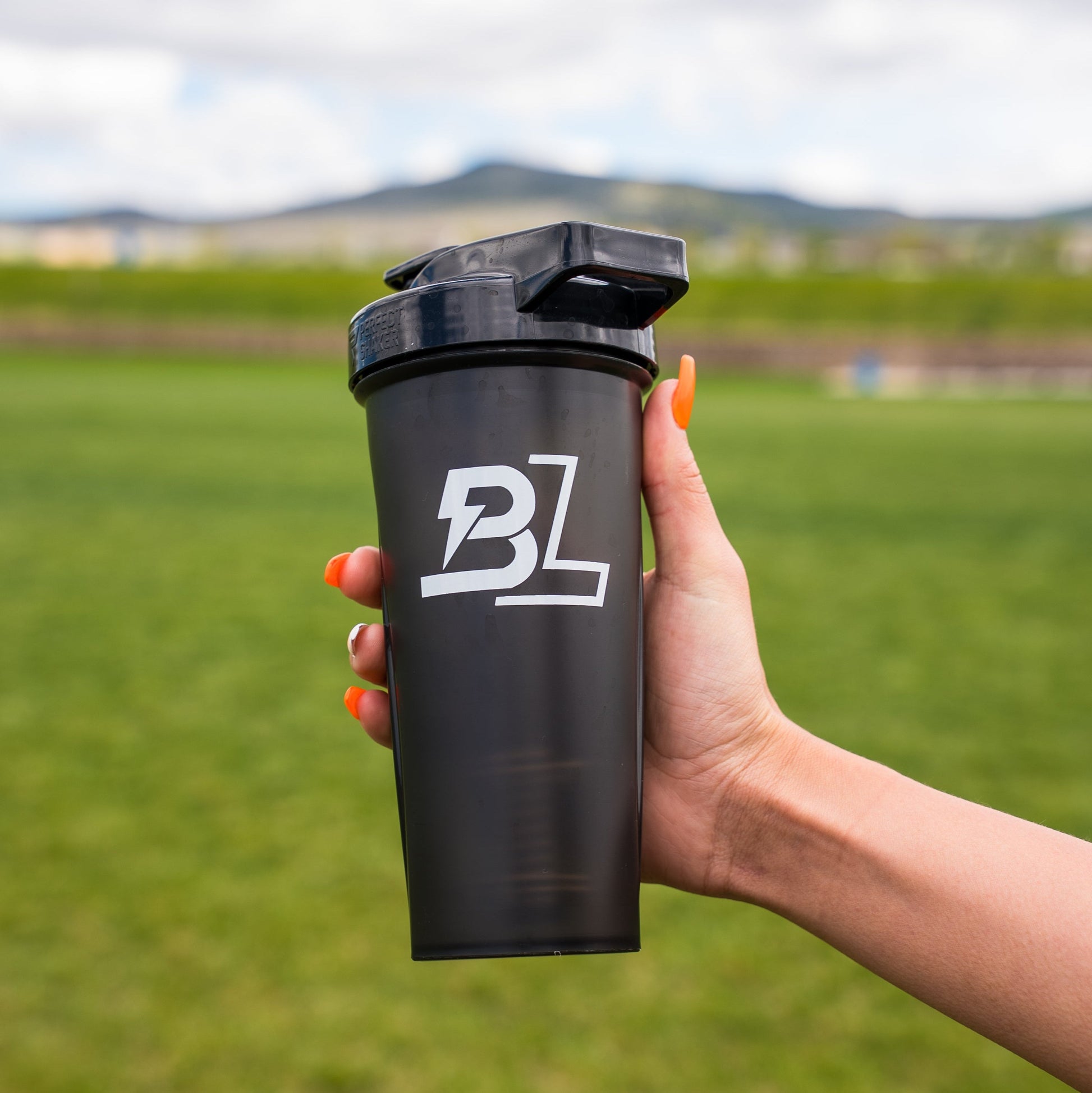 Hand holding a black BLACKLABEL 28 oz protein shaker bottle with white logo in an outdoor grassy field, showcasing a reusable shaker cup for mixing protein shakes, pre-workout, or hydration drinks.