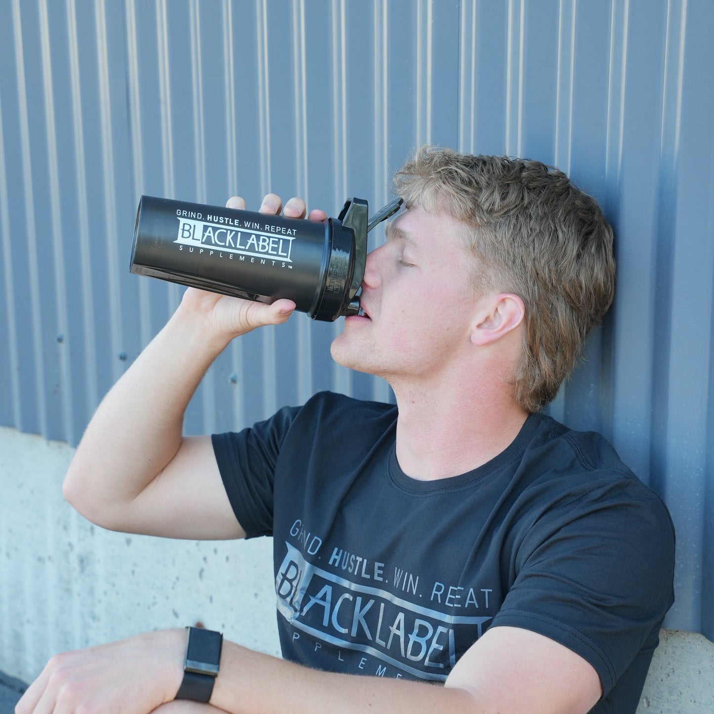 Person drinking from a BLACKLABEL 28 oz shaker bottle while resting after a workout, highlighting a reusable protein shaker cup for mixing protein shakes, hydration, and fitness supplements.