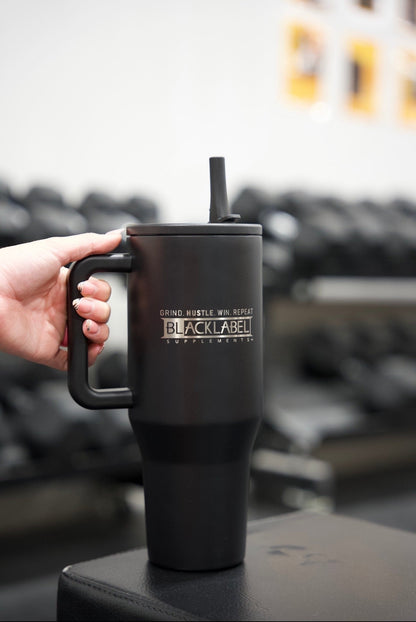 Hand holding a matte black BLACKLABEL 40 oz insulated traveler tumbler with handle and straw in a gym, designed for convenient hydration during workouts, travel, or everyday use.