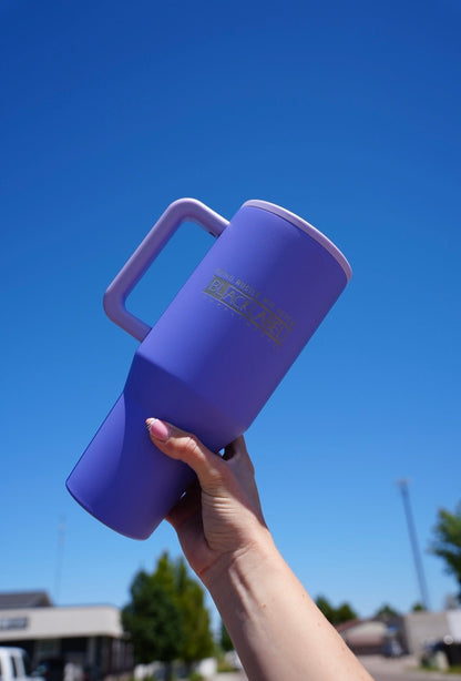 Hand holding a purple BLACKLABEL 40 oz insulated traveler tumbler with ergonomic handle against a clear blue sky, featuring a durable matte finish for hydration at the gym, work, travel, or everyday use.