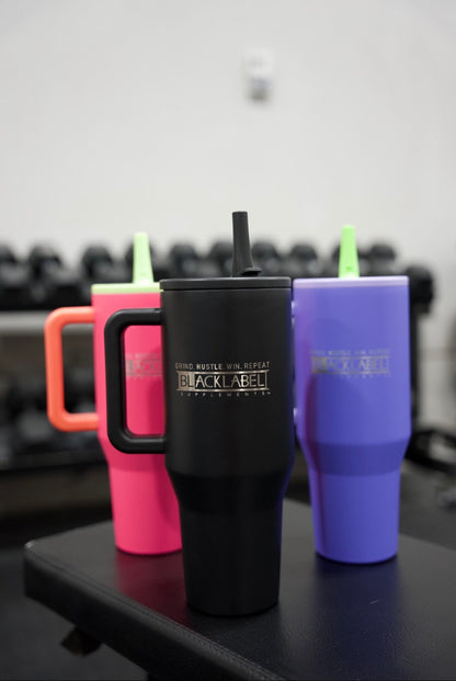 Matte black, purple, and pink BLACKLABEL 40 oz insulated traveler tumblers with handles and straws displayed together in a gym, showcasing multiple color options for hydration on the go.