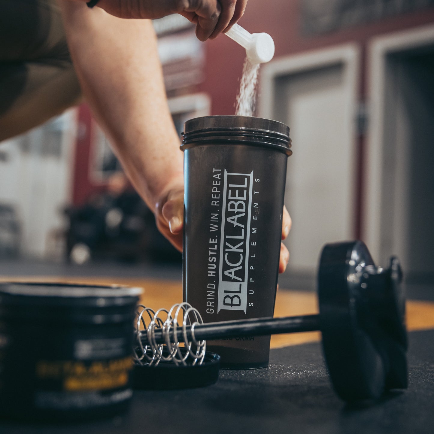 Person adding powdered supplement to a BLACKLABEL protein shaker bottle on a gym floor with a whisk mixer, dumbbell, and supplement container nearby, showcasing pre-workout or protein preparation before training.
