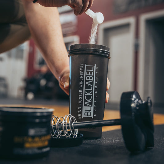 Person adding powdered supplement to a BLACKLABEL protein shaker bottle on a gym floor with a whisk mixer, dumbbell, and supplement container nearby, showcasing pre-workout or protein preparation before training.