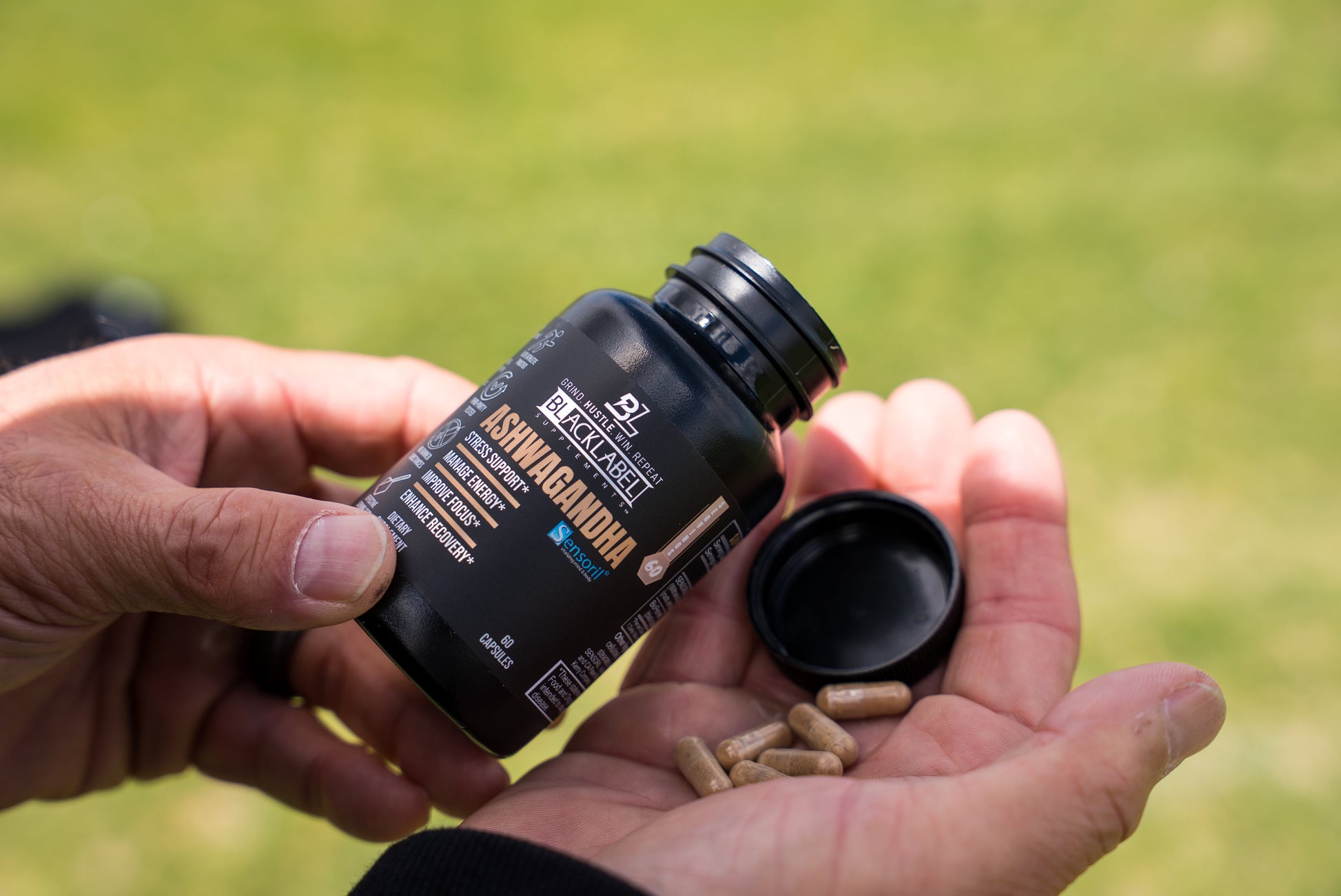 Close-up of a person pouring BlackLabel Ashwagandha Stress Support capsules into their hand, with the supplement bottle and capsules in focus against a softly blurred outdoor background.