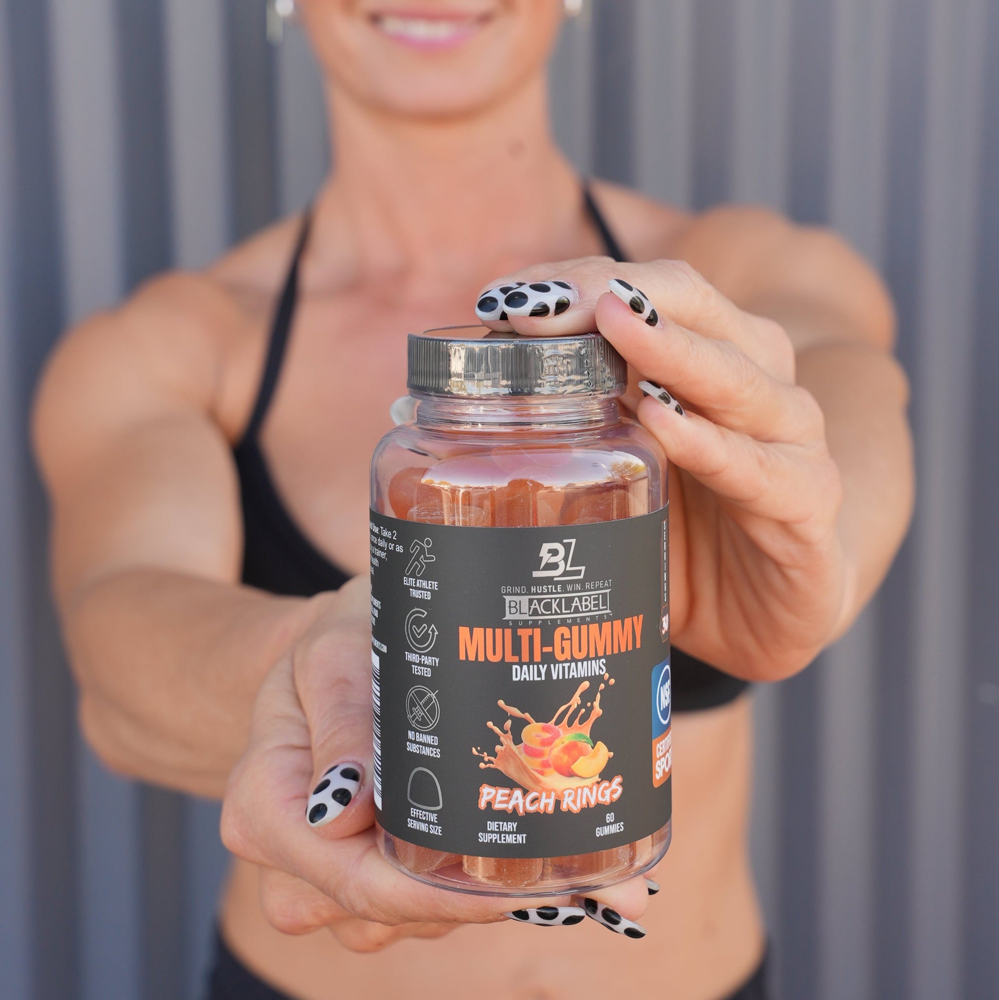 Fitness enthusiast holding a bottle of BlackLabel Multi-Gummy Daily Vitamins in Peach Rings flavor toward the camera with a blurred gym-style background.
