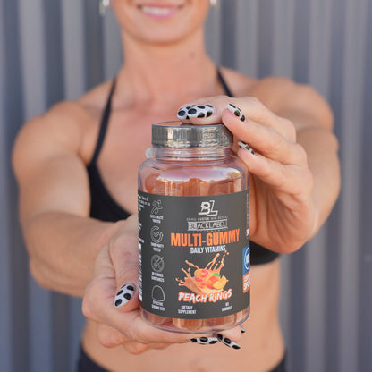Fitness enthusiast holding a bottle of BlackLabel Multi-Gummy Daily Vitamins in Peach Rings flavor toward the camera with a blurred gym-style background.
