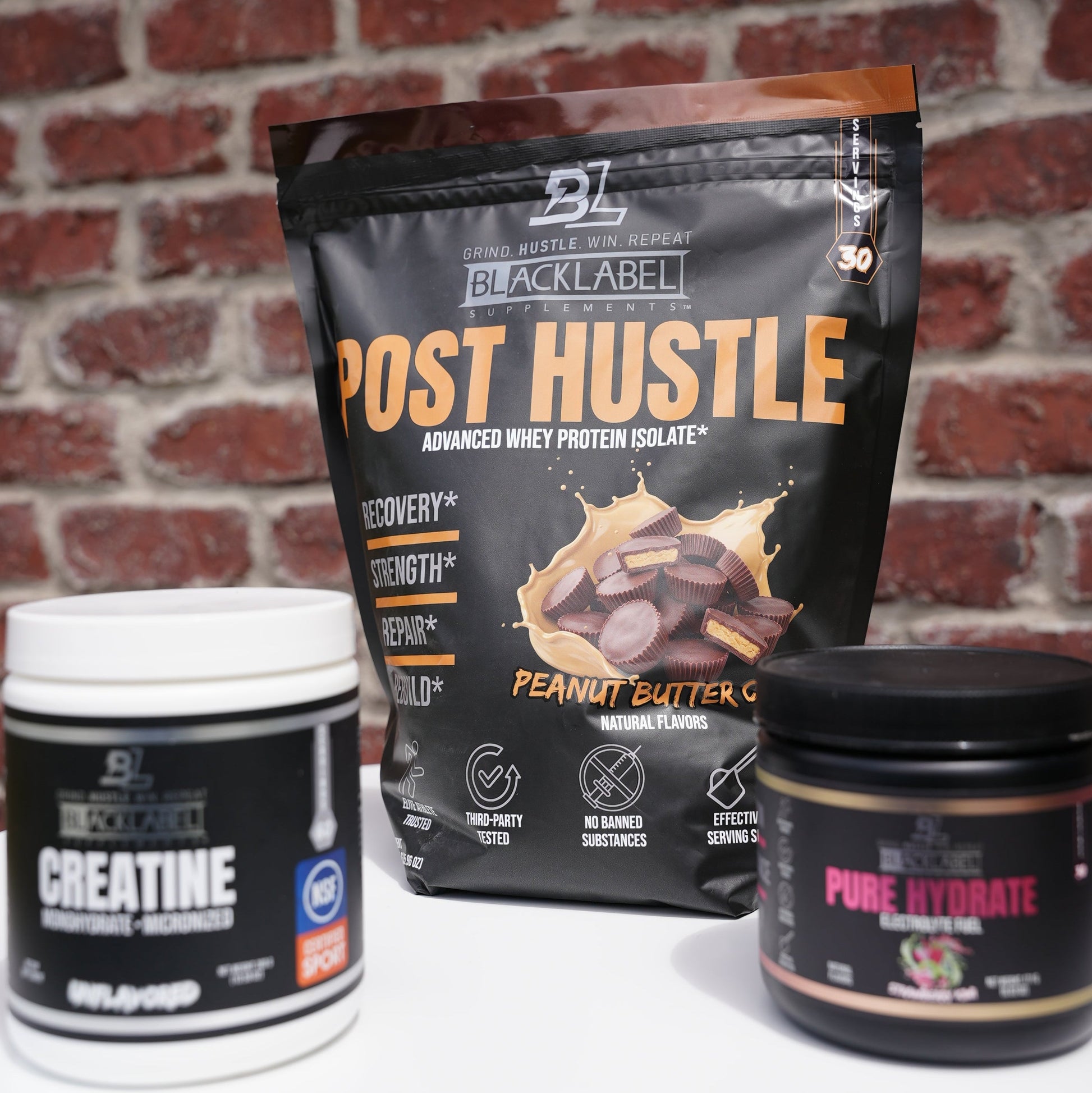 BlackLabel post-workout recovery supplements displayed on a white surface in front of a brick wall, featuring Post Hustle whey protein isolate, Creatine Monohydrate, and Pure Hydrate electrolyte drink mix to support muscle recovery, hydration, strength, and performance.