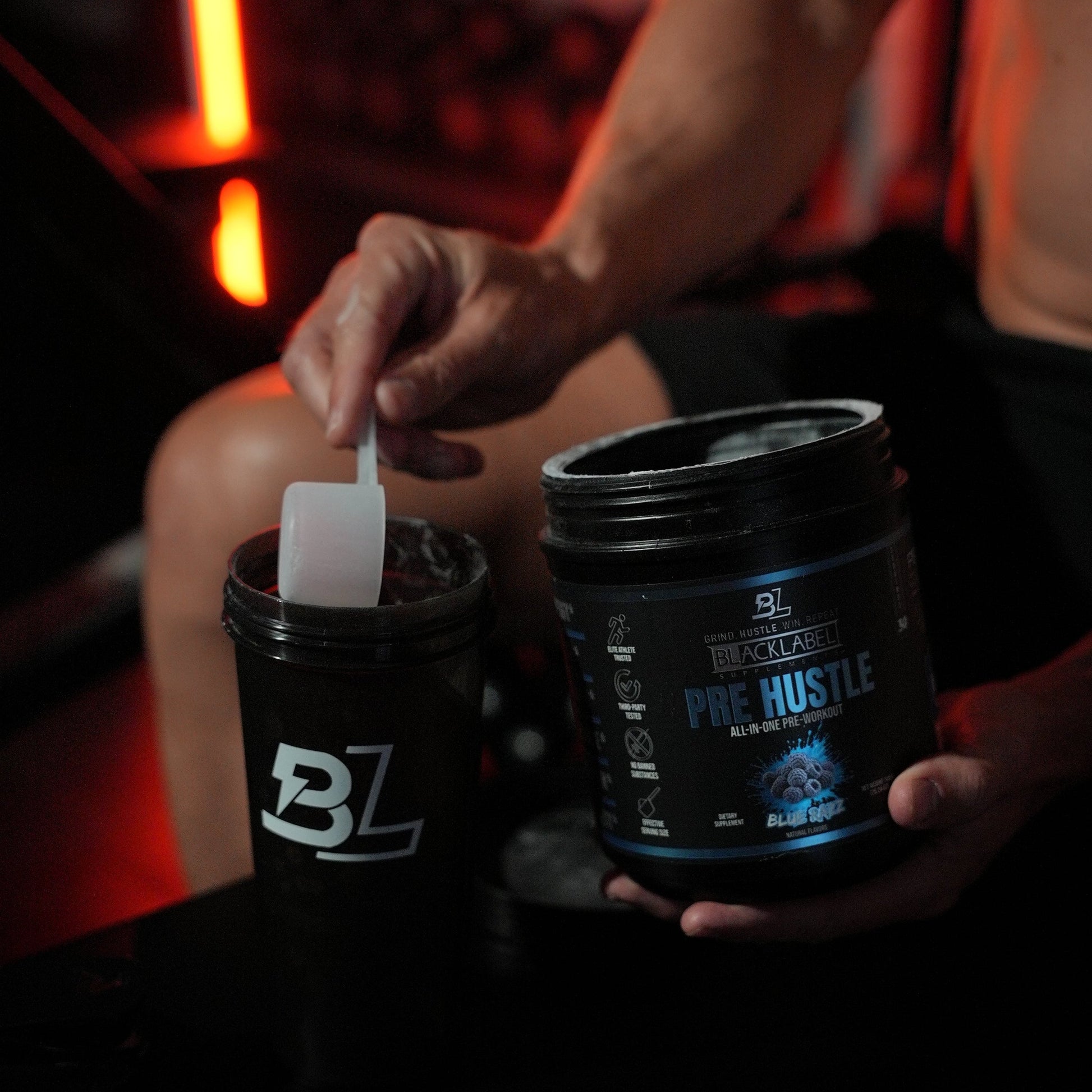 Hand scooping BlackLabel Pre Hustle Blue Razz pre-workout powder into a branded shaker bottle in a gym, demonstrating quick pre-workout drink preparation before exercise.