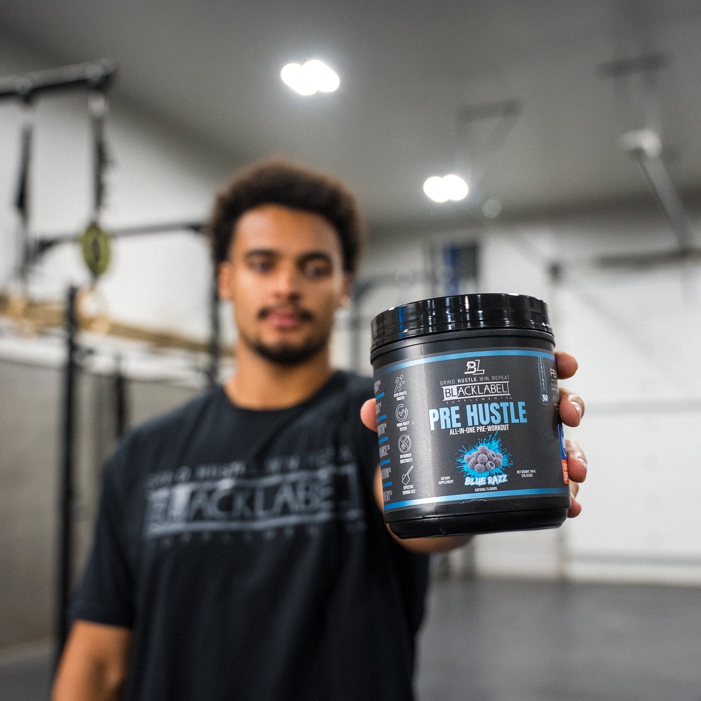 Athlete holding a container of BlackLabel Pre Hustle Blue Razz pre-workout supplement in a gym, showcasing the product before a strength training workout.