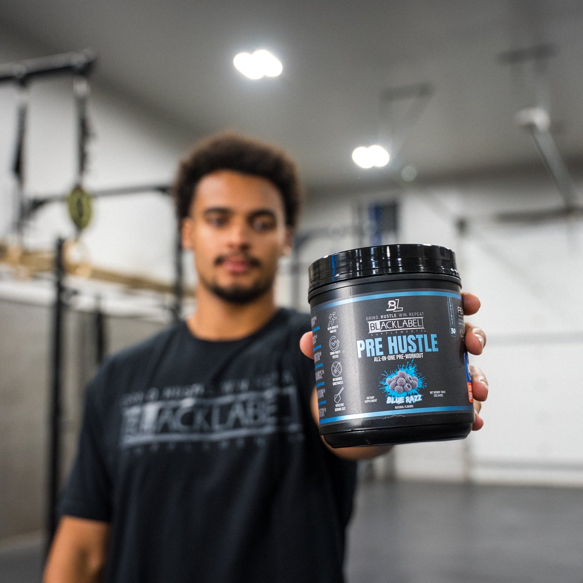 Athlete holding a container of BlackLabel Pre Hustle Blue Razz pre-workout supplement in a gym, showcasing the product before a strength training workout.