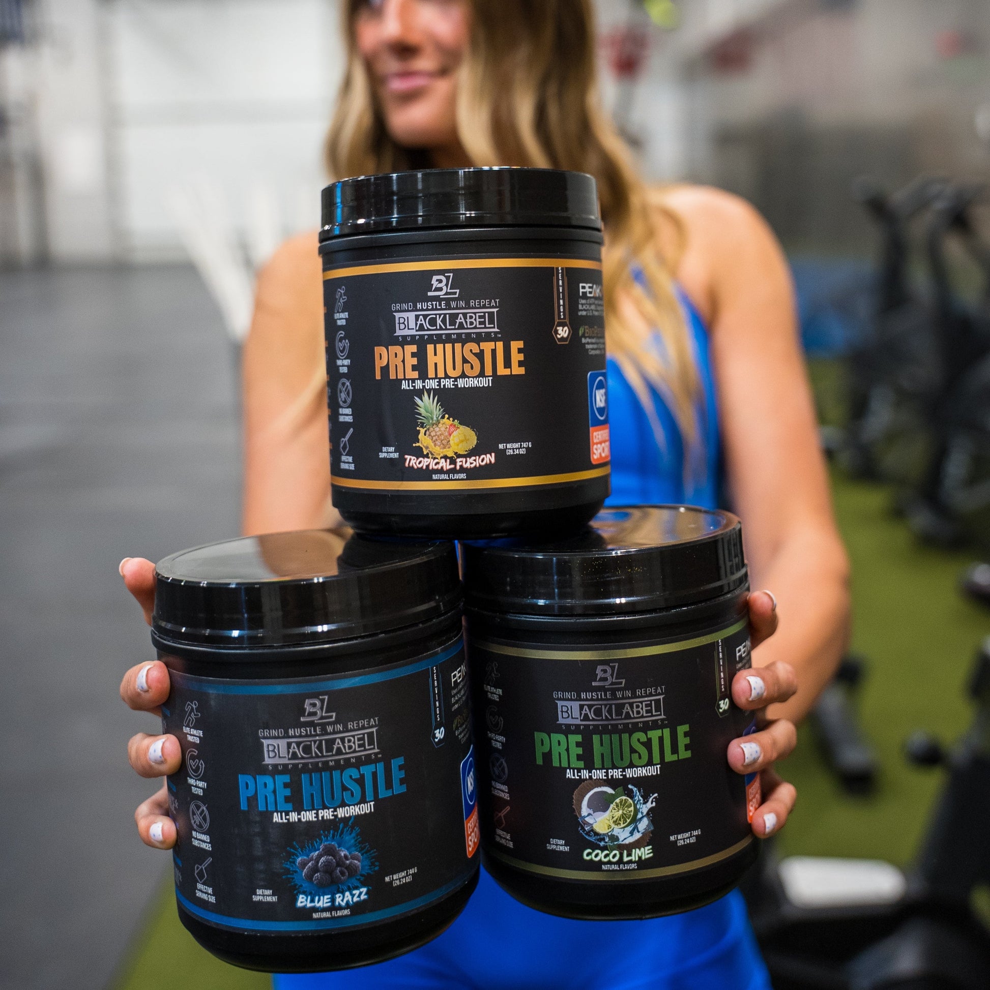 Fitness athlete holding three BlackLabel Pre Hustle pre-workout supplements in Blue Razz, Coco Lime, and Tropical Fusion flavors inside a gym, showcasing multiple flavor options for active lifestyles.