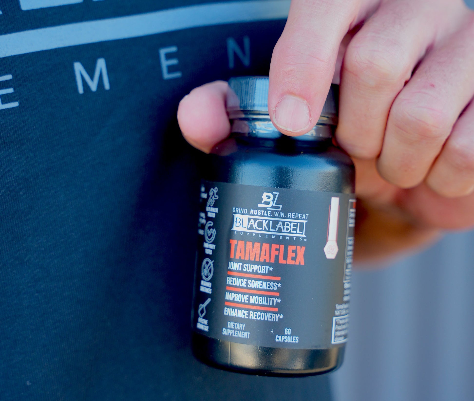 Close-up of a person holding a bottle of BlackLabel TamaFlex Joint Support supplement, with the product label in focus against a softly blurred fitness background.