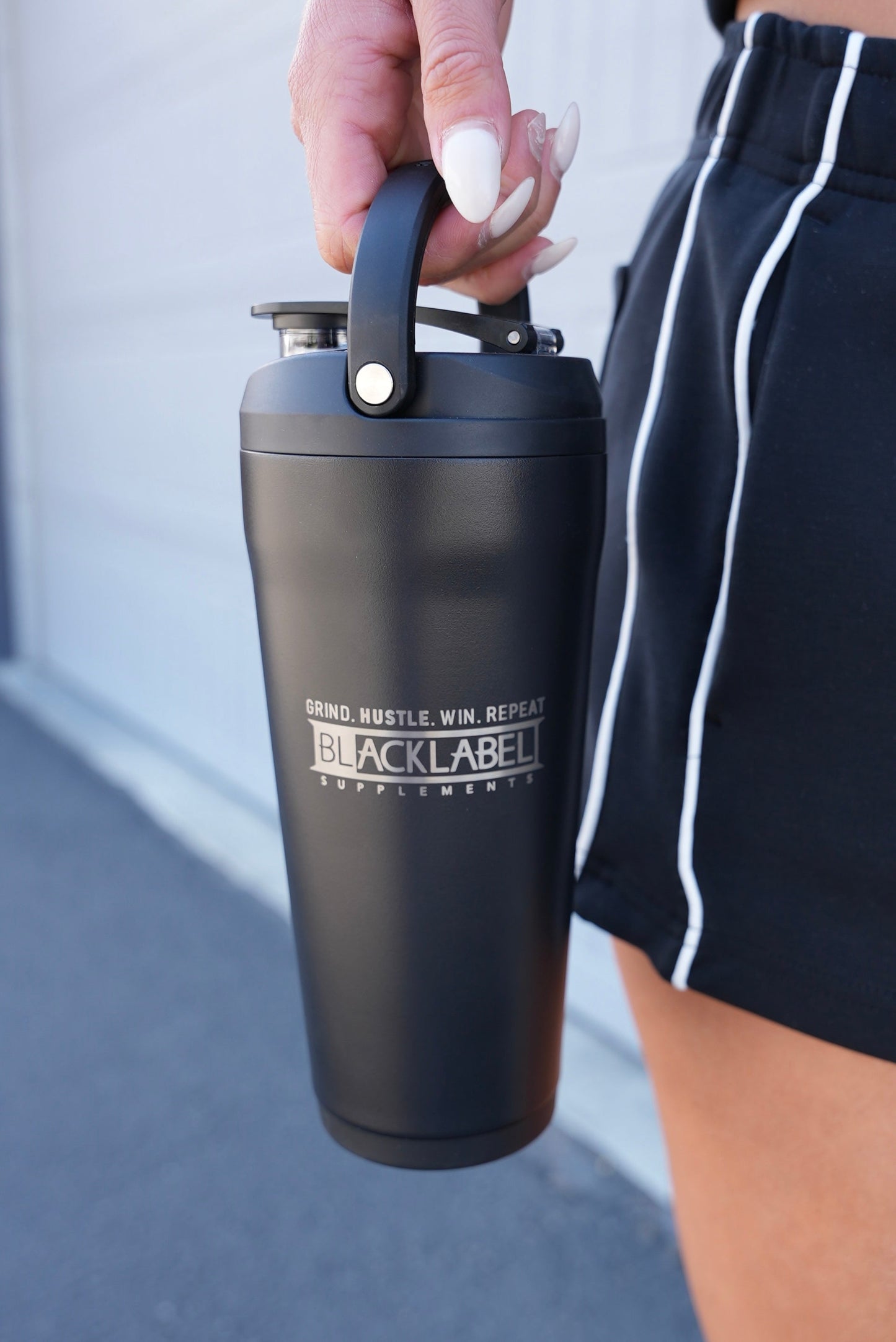 Black BLACKLABEL 24 oz insulated shaker bottle with built-in carry handle, held by hand outdoors, ideal for protein shakes, hydration, workouts, travel, and everyday use.