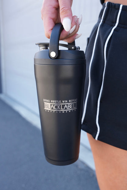 Black BLACKLABEL 24 oz insulated shaker bottle with built-in carry handle, held by hand outdoors, ideal for protein shakes, hydration, workouts, travel, and everyday use.