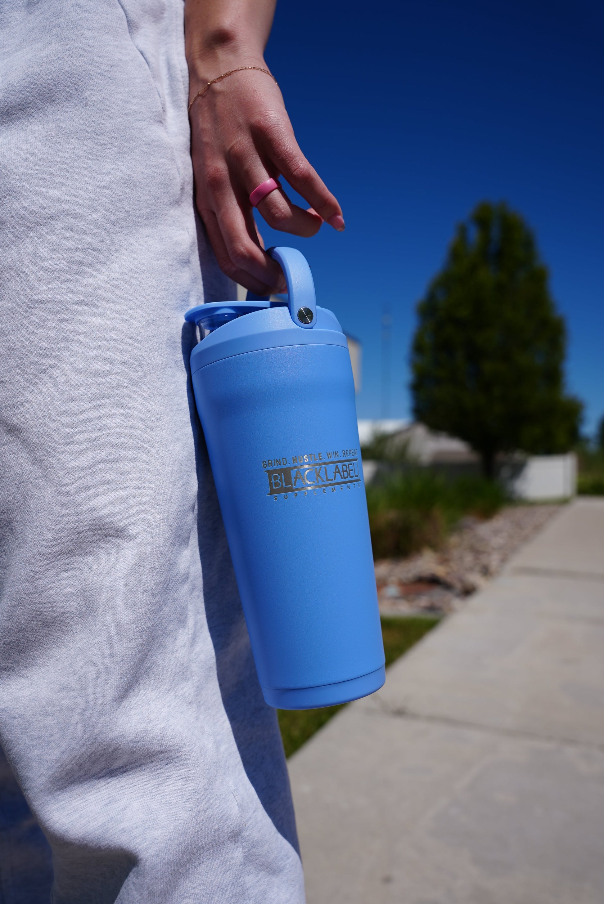 Person carrying a blue BLACKLABEL 24 oz insulated stainless steel shaker bottle with a built-in handle and leak-resistant flip lid, designed for protein shakes, hydration, workouts, and everyday use.