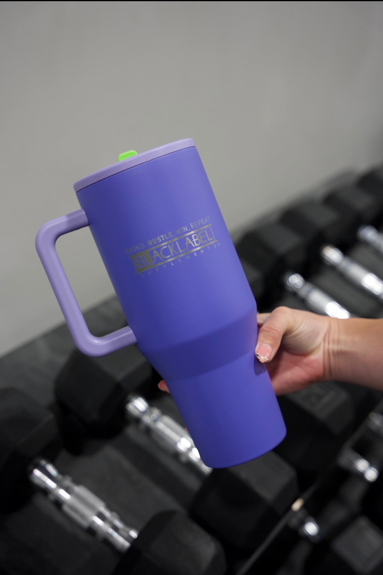 Hand holding a purple BLACKLABEL 40 oz insulated traveler tumbler with handle and straw above gym dumbbells, ideal for staying hydrated during workouts, travel, or everyday use.