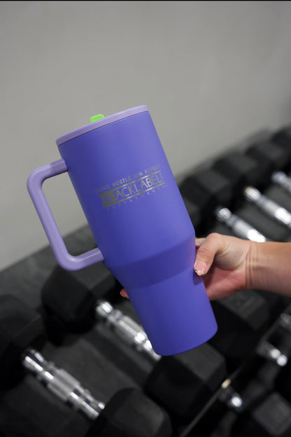 Hand holding a purple BLACKLABEL 40 oz insulated traveler tumbler with handle and straw above gym dumbbells, ideal for staying hydrated during workouts, travel, or everyday use.