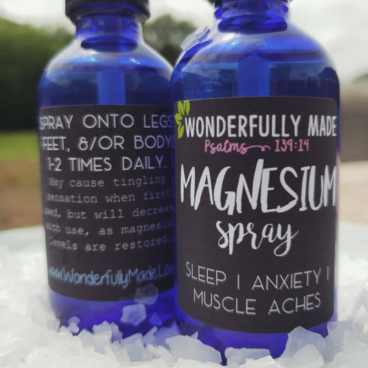 Two cobalt blue glass bottles of topical magnesium spray with wellness-themed labels displayed on a bed of mineral flakes, highlighting recyclable packaging and a fine-mist spray design.