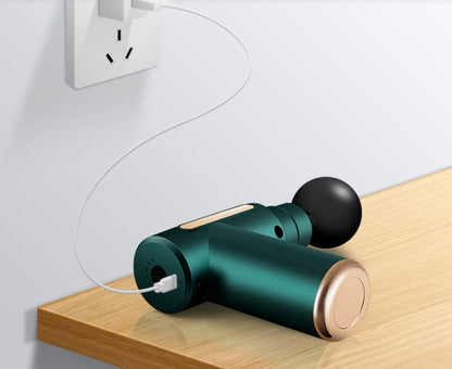 Green cordless percussion massage gun connected to a USB charging cable on a wooden tabletop, featuring a rechargeable battery and round massage head for portable muscle massage and everyday relaxation.