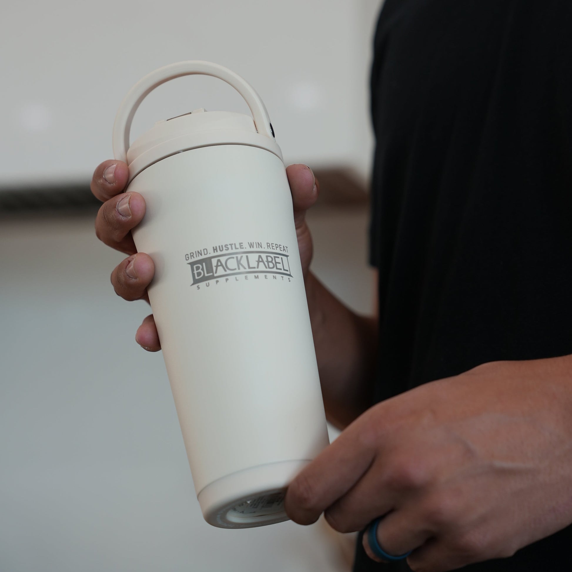 Person holding a white BLACKLABEL 24 oz insulated stainless steel shaker bottle with a built-in carry handle, designed for protein shakes, hydration, workouts, travel, and everyday use.