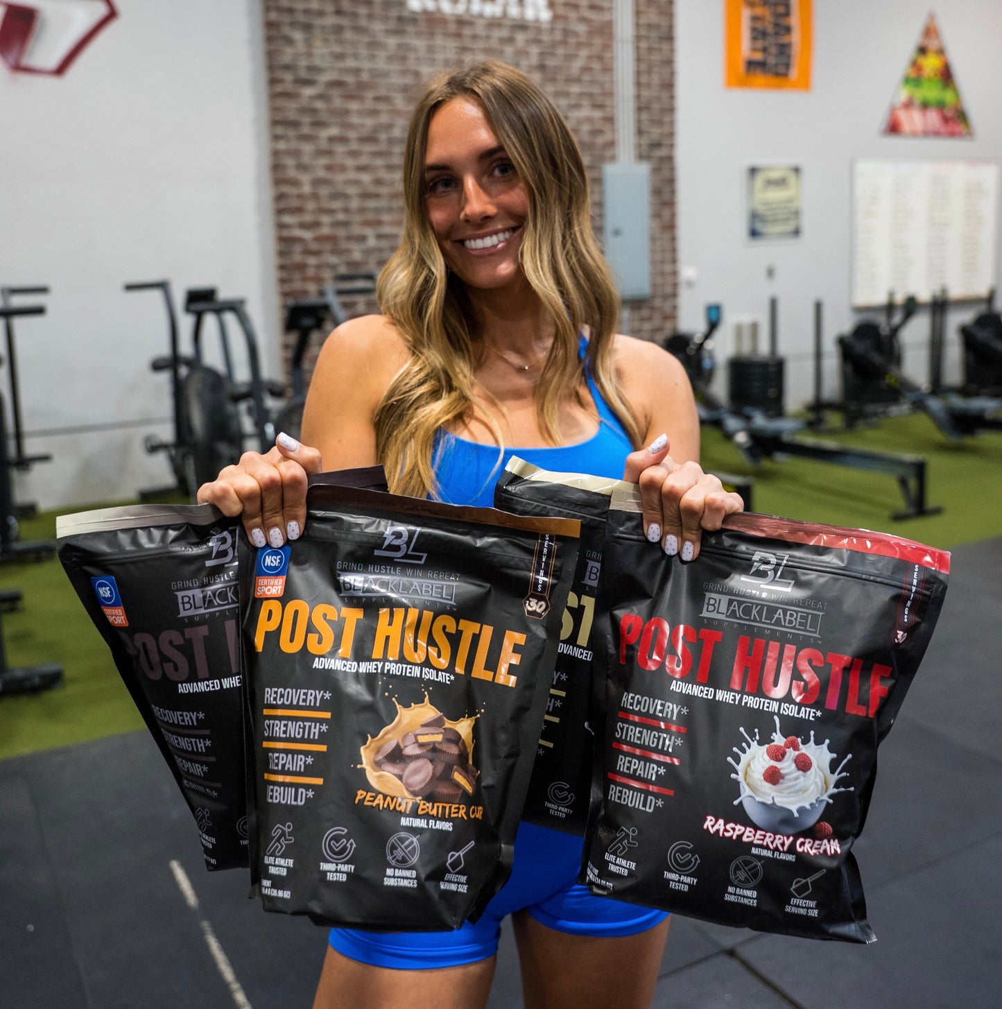 Woman holding Black Label Post Hustle protein powder packages in a gym setting