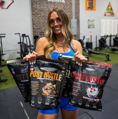 Woman holding Black Label Post Hustle protein powder packages in a gym setting